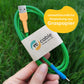 recable uses an environmentally friendly band made from grass paper. Representation: In one hand is a finished green, blue and orange USB-A to USB-C charging cable. The band shows the recable logo and the inscription "charge your life". In the background there is a meadow.